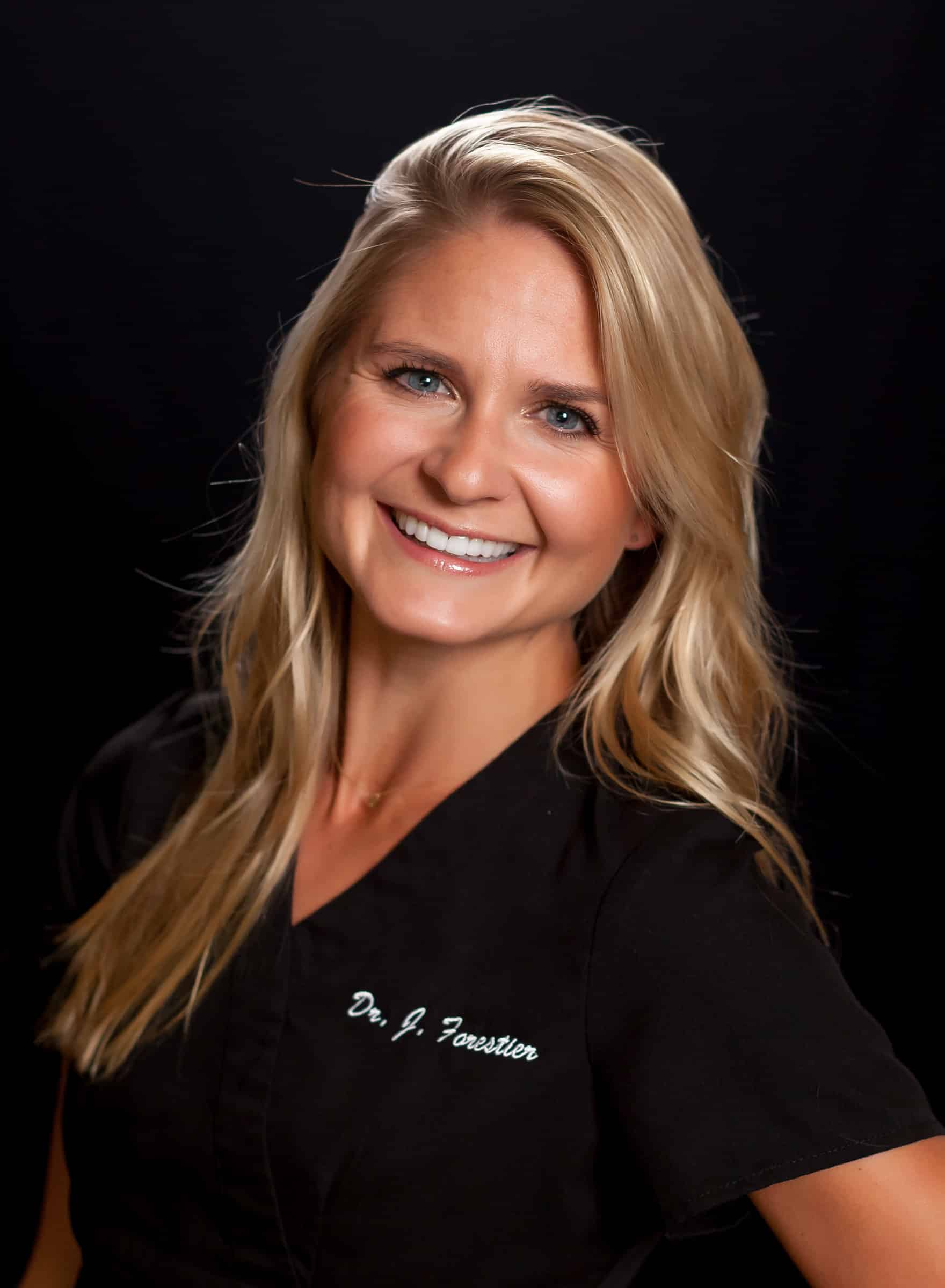 Head-and-shoulders portrait of a smiling blonde woman with blue eyes, against a black background, wearing a dark top with white cursive text on the chest area.